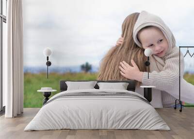 Happy family mom woman with little boy hugging at weekend. Hug, background green grass. Warm casual wear, hooded sweater. Caring support love and nurturing tenderness. Free space for text. Wall mural