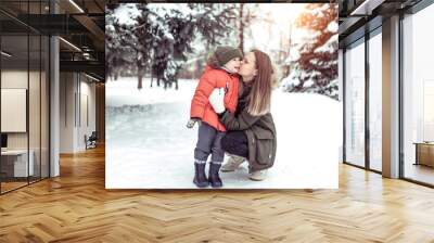 Young woman mother kisses her child, a boy of 3 years. In winter, outdoors in the park, background of snow trees spruce. The concept of a walk on weekends holidays. Happy family on a walk play. Wall mural