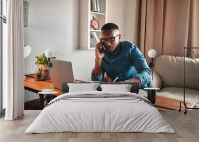 Young entrepreneur on his phone while taking notes at his desk. Professional man working remote from home with technology. African American male has a business meeting on an audio call. Wall mural