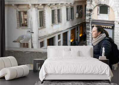 A woman wearing a puffer and a scarf taking pictures of a seagull at Rialto Bridge in Venice; Italy Wall mural