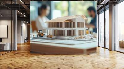 Model of a house and construction equipment on a table, with two people working facing each other behind it, blurred background Wall mural