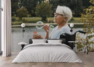 senior gray haired disabled woman in the wheelchair blowing soap bubbles near the river. High quality photo Wall mural