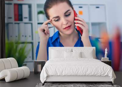 Young serious Caucasian businesswoman sitting at desk in office, speaking on the phone. Wall mural