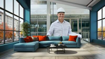 The boss with the plan. Portrait of a cheerful professional male architect looking at the camera while holding blueprints inside a building. Wall mural