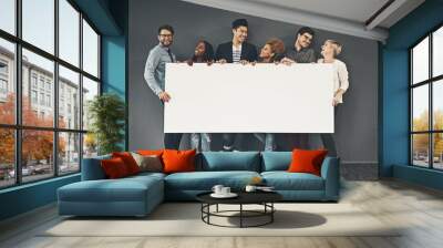 A copyspace of a diverse group of young, happy and smiling professional business people holding a blank white billboard. A multiracial team of men and women against a dark wall with copy space Wall mural