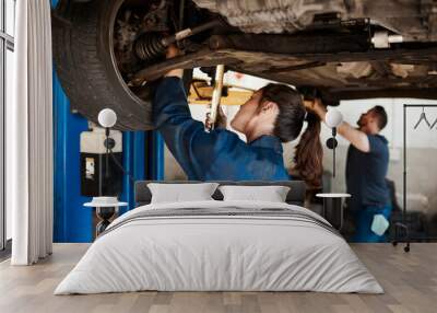 Mechanic, woman and man under car in workshop for auto repair, service and inspection for safety. Engineering, partnership and teamwork for vehicle maintenance with tools, parts or industry at garage Wall mural