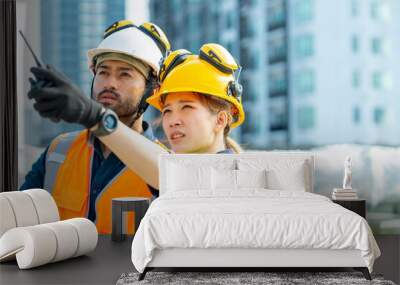 Asian man and woman teamwork engineer working on digital tablet and speaking on two way radios at construction site building rooftop. Architecture inspector inspect building exterior structure system. Wall mural