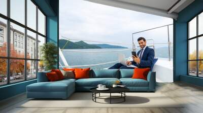 Caucasian businessman working on laptop computer and talking on mobile phone for global corporate business while travel on luxury private catamaran boat yacht sailing in the ocean on summer vacation. Wall mural