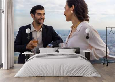 Caucasian couple celebrating at luxury skyscraper rooftop bar at summer sunset. Confident man and woman couple enjoy city lifestyle having dinner and cocktail drink at outdoor restaurant in evening. Wall mural