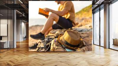 Caucasian man traveler reading a book during resting on coastline hill at sunset. Handsome guy enjoy outdoor lifestyle hiking on island mountain on summer holiday vacation. Solo travel concept. Wall mural