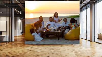 Group of multiethnic family friends enjoy dinner party together on the beach at sunset. Diverse family with child girl, adult and senior couple relax and having fun together on summer holiday vacation Wall mural