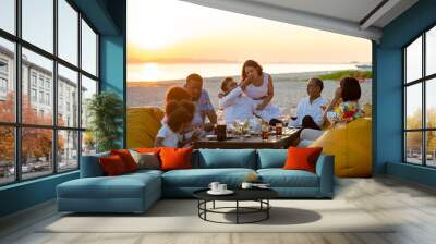 Group of multiethnic family friends enjoy dinner party together on the beach at sunset. Diverse family with child girl, adult and senior couple relax and having fun together on summer holiday vacation Wall mural