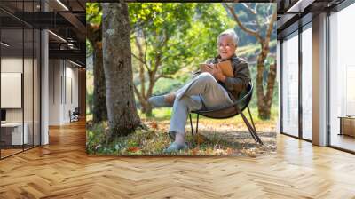 Happy Asian senior man sitting on outdoor chair in nature public park and reading a book. Healthy elderly retired man relax and enjoy outdoor lifestyle leisure activity on summer holiday vacation. Wall mural