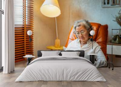 Happy Asian senior mature woman wearing glasses relaxing on sofa in living room and reading a book. Elderly retired grandmother enjoy indoor lifestyle with leisure activity and hobbies at home. Wall mural