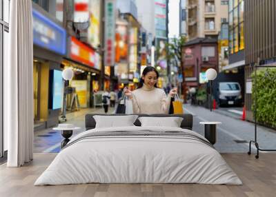 Happy Asian woman holding shopping bag walking at Shibuya district, Tokyo, Japan in evening. Attractive girl enjoy and fun outdoor lifestyle travel urban city street and shopping on holiday vacation. Wall mural