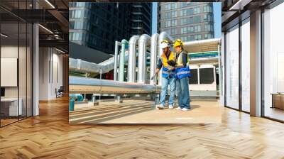 Professional Asian man and woman teamwork engineer in safety uniform working at outdoor construction site rooftop. Industrial technician worker maintenance checking building exterior plumbing systems. Wall mural