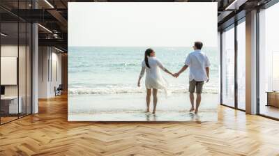 Rear view of Happy young Asian couple family man and woman holding hands together and walking on the beach in summertime. Healthy people enjoying relax lifestyle in romantic summer holiday vacation. Wall mural