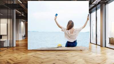 Rear view of young beautiful asian woman girl sitting on rocky seacoast beach with holding smartphone and credit card and outstretched her arms in the air for success online shopping with e-banking Wall mural