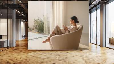 A woman sits in a cozy chair, holding a cup of coffee, gazing out the window and enjoying a peaceful moment of relaxation and contemplation. Wall mural