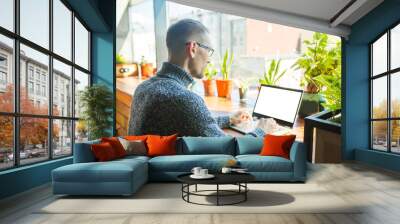 Concentrated young businessman in eyewear sitting and looking at laptop computer screen near large window. Working distantly online, work on project and freelance concept. Wall mural