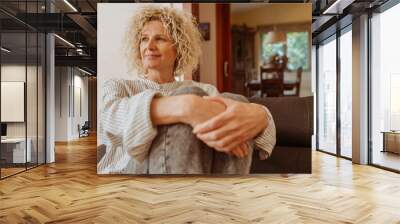 Beautiful smiling relaxed middle aged woman relaxing on sofa at home. Stylish middle aged 50s lady with curly hair, sitting on couch holding a pillow in modern living room. Wall mural