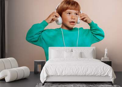 Cute little kid boy 5 years old listen music with headphones, wears cyan sweater isolated on pastel beige background. Childhood lifestyle concept. Mock up copy space. Wall mural