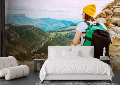 Rear view portrait of a brave young woman wears yellow knit beanie hat and casual clothing, with green travel backpack hiking in mountains outdoor journey. Active traveler lifestyle. Wall mural