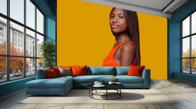 Side view portrait of a young smiling african girl in casual orange dress, posing over yellow background at studio.
 Wall mural