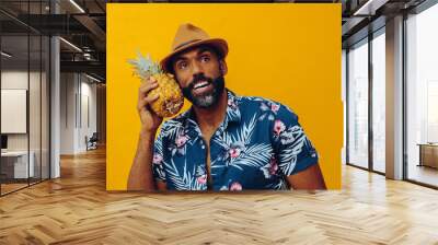 handsome mid adult bearded african american man in Hawaiian shirt posing with pineapple as telephone looking up on yellow background studio Wall mural