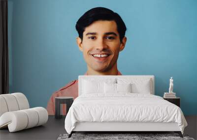 Headshot hispanic latino man black hair smiling handsome young adult wearing pink shirt over blue background looking at camera studio shot Wall mural