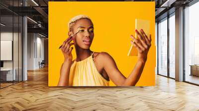 Portrait of a beautiful young woman applying yellow eye shadow make up to her eyes and holding compact mirror Wall mural