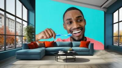 Portrait of a happy young man in pink shirt and great smile brushing teeth with toothbrush and looking to side, isolated on blue Wall mural