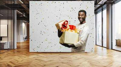 Portrait of a happy young man laughing and holding gold and red gifts as golden confetti are falling, isolated ion a gray studio background Wall mural