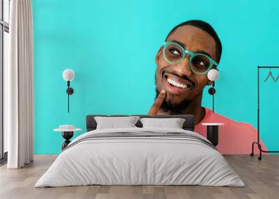 Portrait of a happy young man with glasses smiling, thinking and looking up with finger on chin, against blue studio background Wall mural