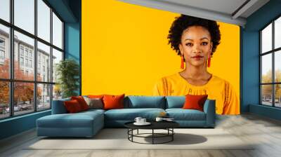 Portrait of a serious young woman with orange tassel beaded earrings on yellow color background Wall mural