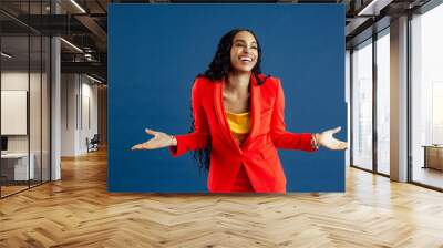 Portrait of a very happy young smiling woman in red business suit with arms out, isolated on blue background Wall mural