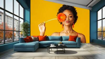 Portrait of a y young woman holding orange Gerbera daisy covering her eye and putting lips sending kiss Wall mural