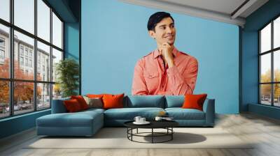 Portrait smiling handsome young adult latino man with arms crossed and hand on chin black hair and pink shirt over blue background looking away at copy space studio shot Wall mural