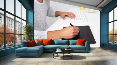 young graphic designer working in his computer in a desktop. He is using a graphic tablet. He is wearing a gray sweater. On the desk there is a cup of coffee and an apple Wall mural