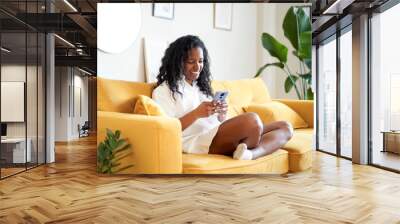 Cheerful Woman using mobile phone at home. African American smiling lady is laying on the sofa. Wall mural