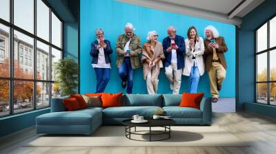 Group of elderly Caucasian laughing together using cell phone standing leaning on blue wall. Happy old friends with gray hair gathered having fun looking mobile outdoor. Mature people enjoying devices Wall mural