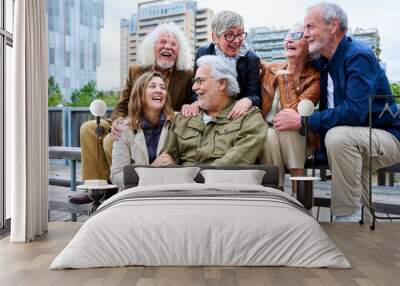 Group of elderly Caucasians laughing together sitting on bench in city. Happy old friends with gray hair gathered having fun outdoors. Lifestyle of cheerful mature people enjoying sunny autumn day Wall mural