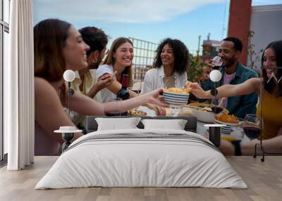 Group of multi-ethnic young friends enjoying meal gathered on rooftop. Happy millennial people having fun drinking red wine and eating celebrating a party event on summer day Wall mural