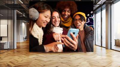 Group of multiracial friends smiling looking and pointing at cell phone outdoor at cold winter night. Four college students enjoying technologies using mobile standing together at amusement park.  Wall mural