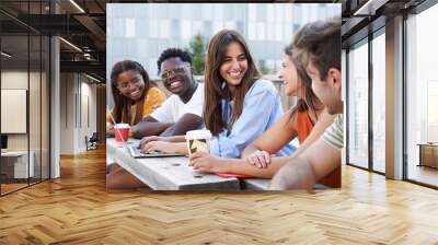 Group of students sitting down in university campus outdoors and smiling. One girl is using a laptop and the others drink coffee. It is a multiracial group of diverse countries. Wall mural