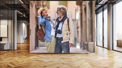 Laughing adult Caucasian couple standing embraced affectionately in tourist street of town. Mature people in love enjoying romantic vacation time. Positive relationships and weekend getaways spring. Wall mural