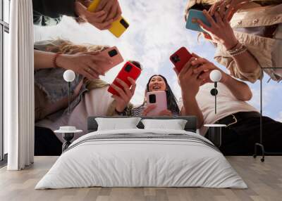 Low angle view of group happy unrecognizable people in circle using colorful mobiles phones together outdoor. Young friends and smiling Asian woman in center holding cells. Gen z technology addiction Wall mural