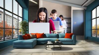 Multiracial group of high school students in the classroom using laptop computers. A smiling Asian girl studies through digital media in new technologies. Wall mural