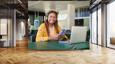 Portrait of a young black girl using headphones a phone and laptop. She looks at the camera and smiling. He is sitting down indoors the college or university campus. Latin people  Wall mural