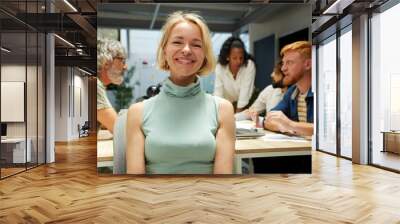 Portrait of an office cheerful Nordic mature woman and looking at camera. Empowered female leader. In the background there is a group of employees kipping at work. It is an indoor multiracial scene. Wall mural